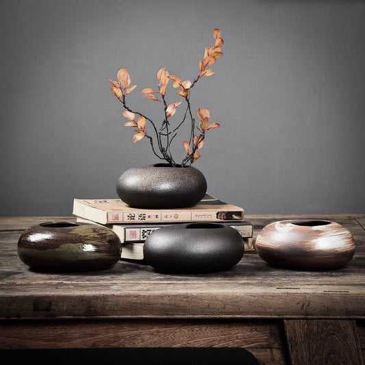 Shan Ying Stoneware Vase Family featuring four round, low-profile ceramic vases in metallic finishes of black, brown, and copper tones, displayed on a rustic wooden surface with books and dried peach-colored flowers arranged in one vase against a gray background